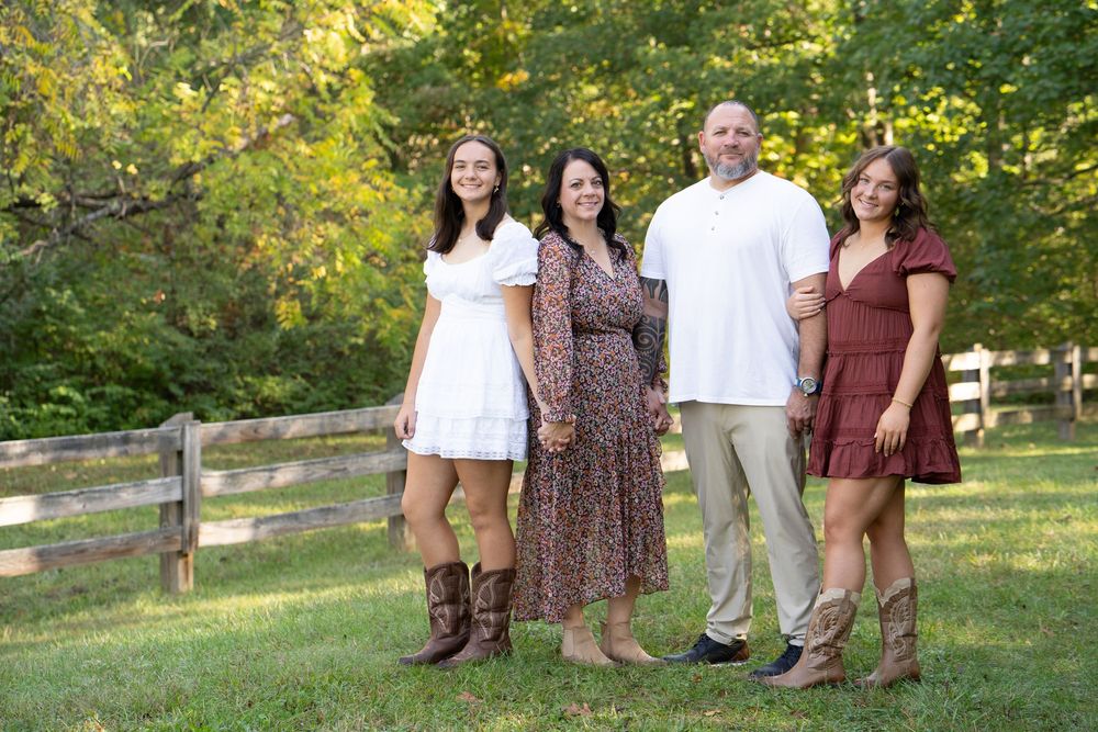 Family of four smiling outdoors on green grass near a wooden fence surrounded by lush trees on a sunny day.