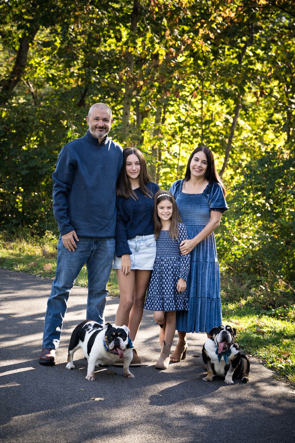 Family of four in blue outfits posing outdoors with two French Bulldogs on a sunny tree-lined path.