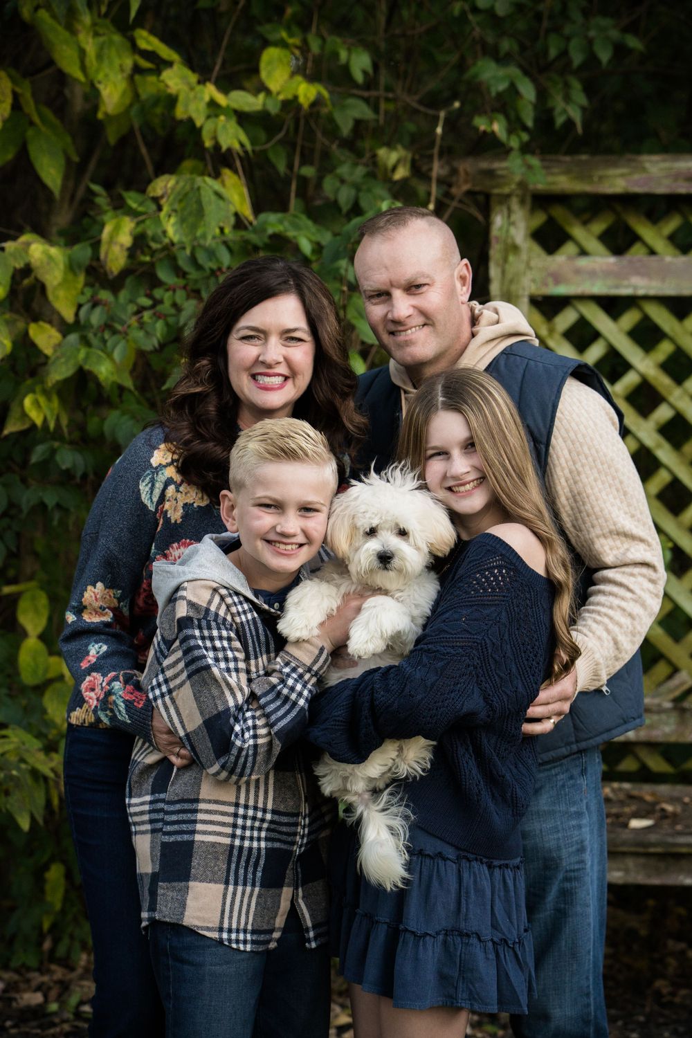 Happy family of four posing outdoors with a white fluffy dog, surrounded by green foliage and a wooden lattice.