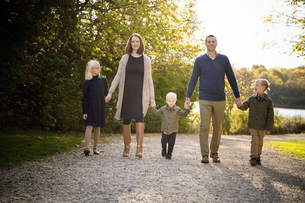 Family of five walking hand-in-hand on a gravel path surrounded by lush green trees during golden hour.