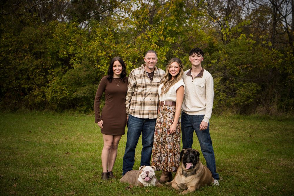 Family of four smiling outdoors with two dogs on green grass, surrounded by autumn trees.