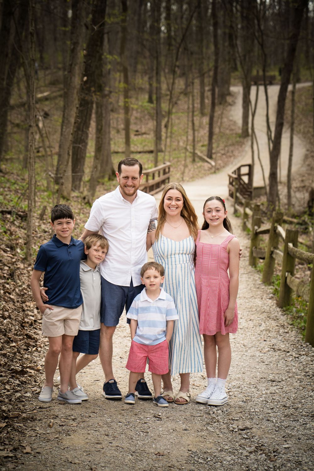 Family of six posing on a wooded trail, parents with four children dressed in casual summer outfits, surrounded by trees.