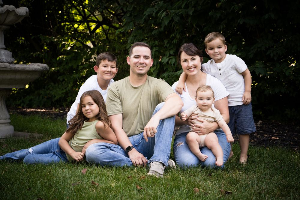 Happy family of six smiling outdoors on green grass, parents with four children posing for a portrait in a lush garden.