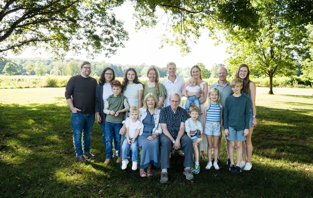 Large multigenerational family posing together outdoors on green grass under shady trees on a sunny day.