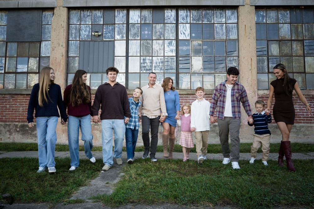 Large family group holding hands in a line outdoors in front of a brick building with large windows.