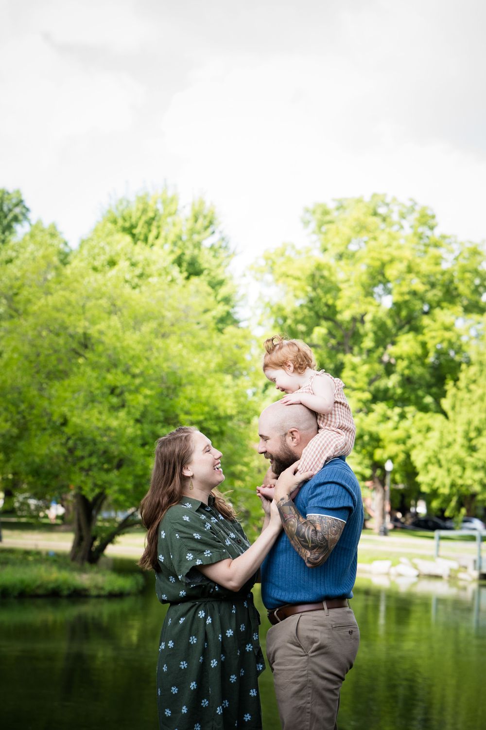 Family of three posing outdoors by a pond, father carrying toddler on shoulders while mother smiles, surrounded by green trees.