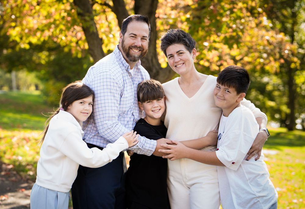 Happy family of five smiling together outdoors in a park with colorful autumn foliage in the background.
