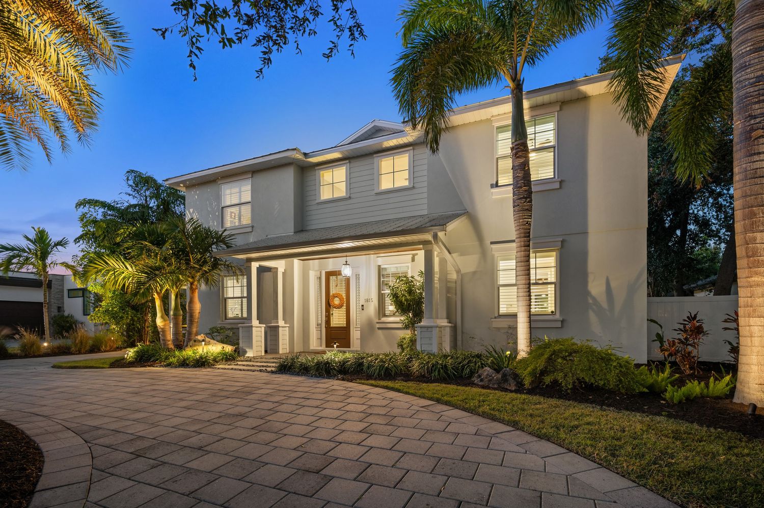 Elegant two-story beige home with palm trees and curved driveway illuminated at dusk.