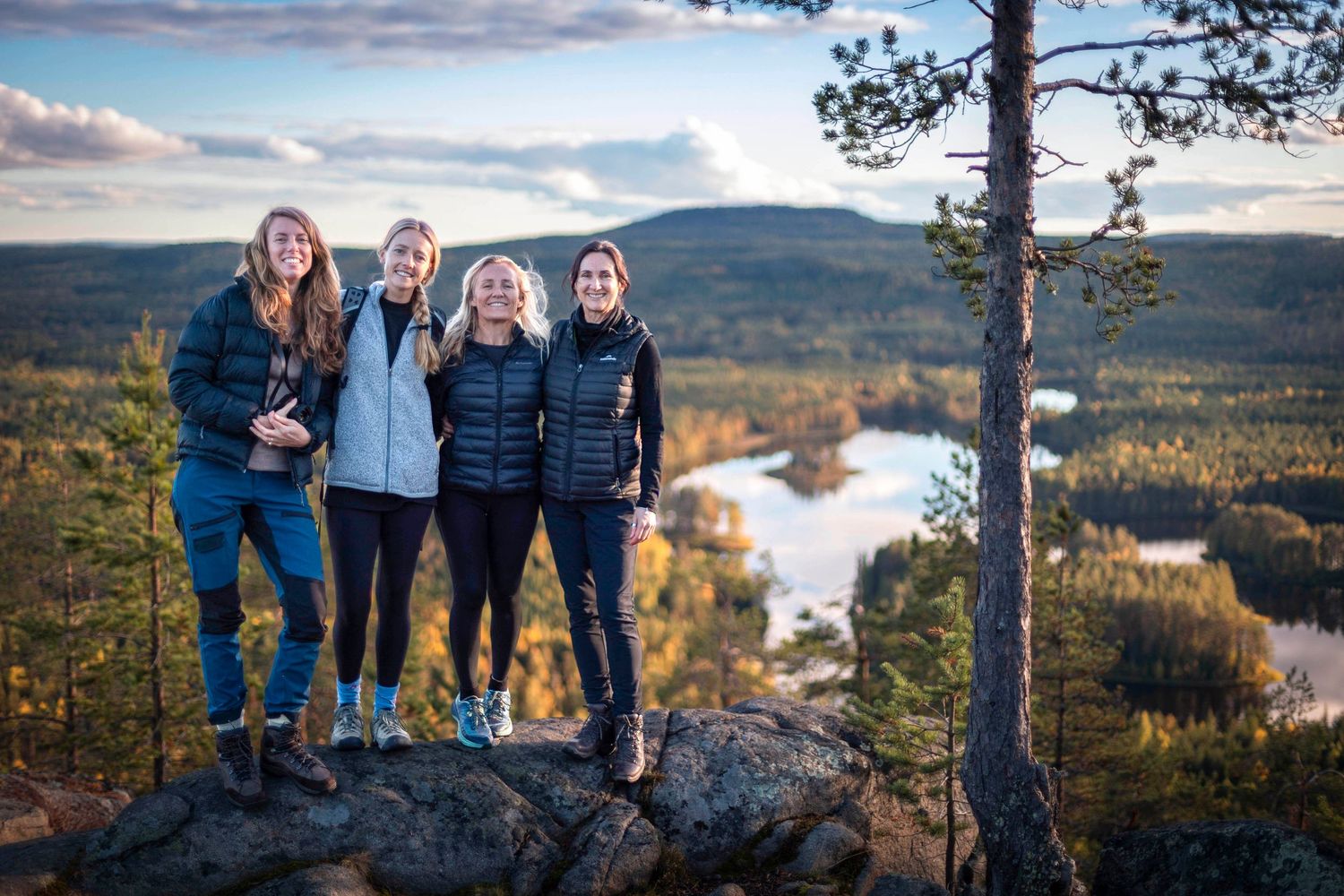 Four women posing on rocky hilltop overlooking scenic autumn forest lake landscape at sunset.