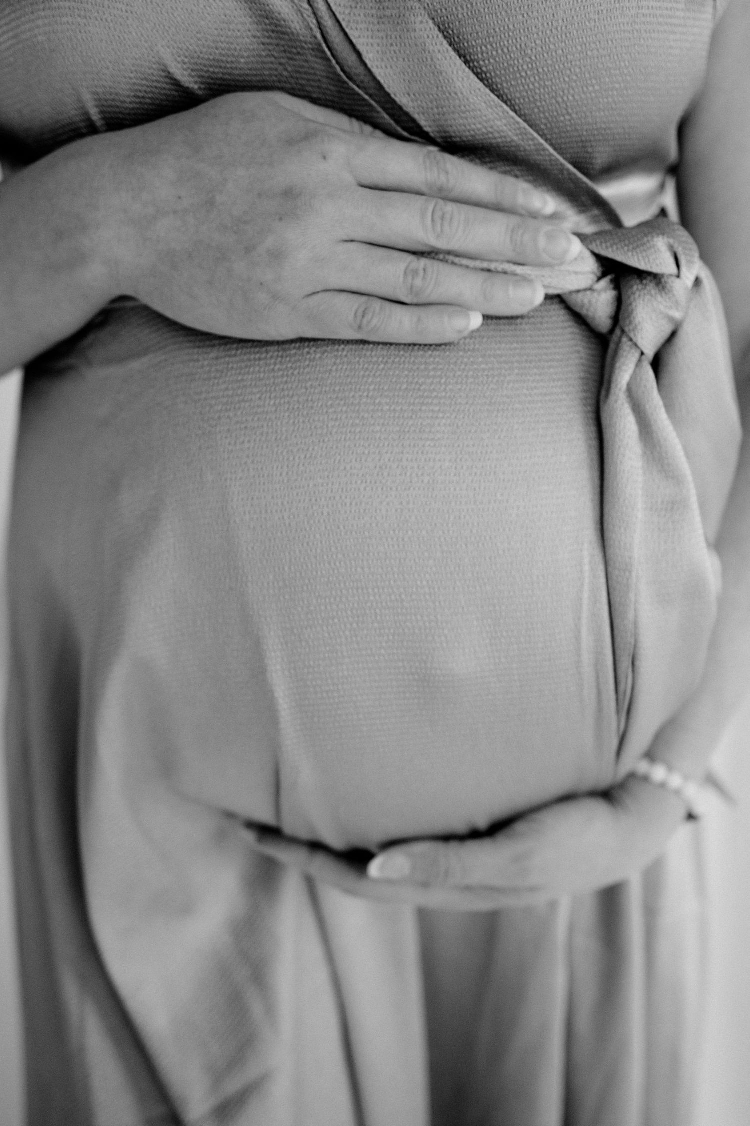 Black and white photo showing hands cradling a pregnant belly through soft flowing fabric in an intimate maternity pose.