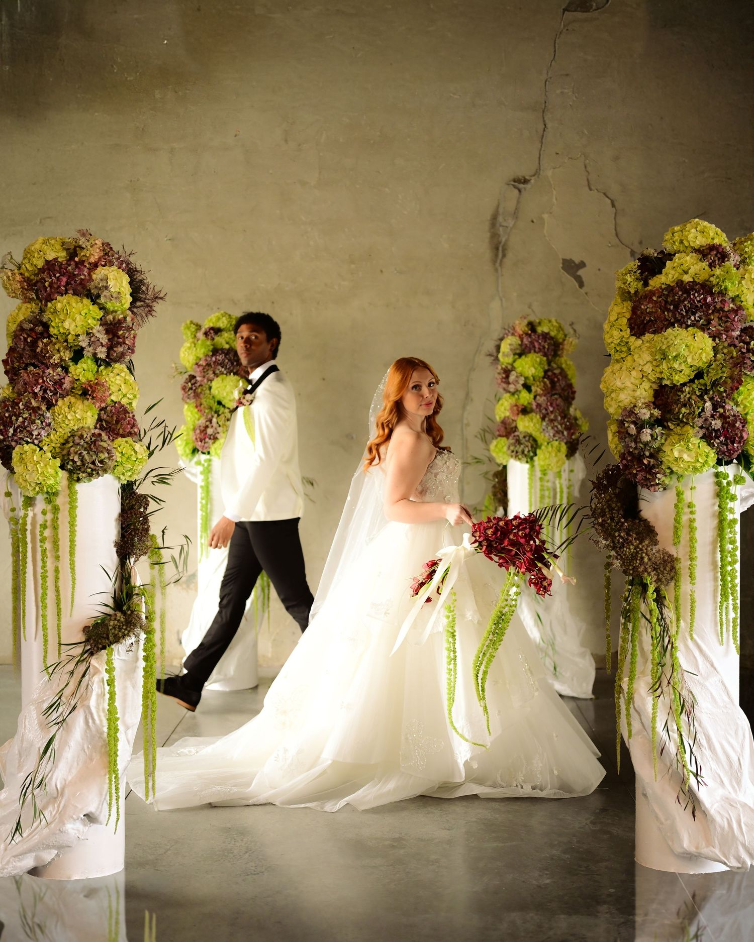Bride and groom walk between tall floral arrangements of green and burgundy blooms on white pedestals in elegant wedding ceremony.