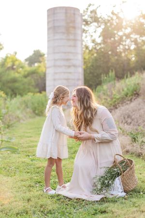Mother and daughter in white dresses share a tender nose-to-nose moment in a sunlit field near a rustic silo.
