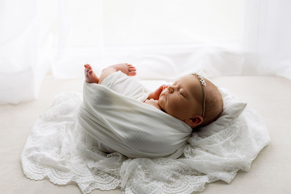 Newborn baby sleeping peacefully wrapped in white swaddle on lace blanket in soft natural light.