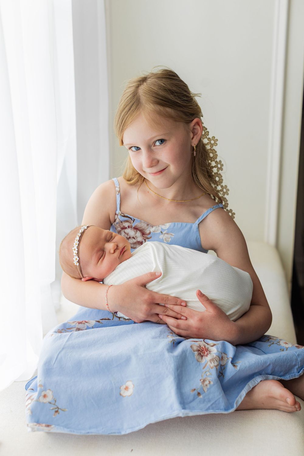 Young girl in blue floral dress holding a swaddled newborn baby while sitting by a bright window.