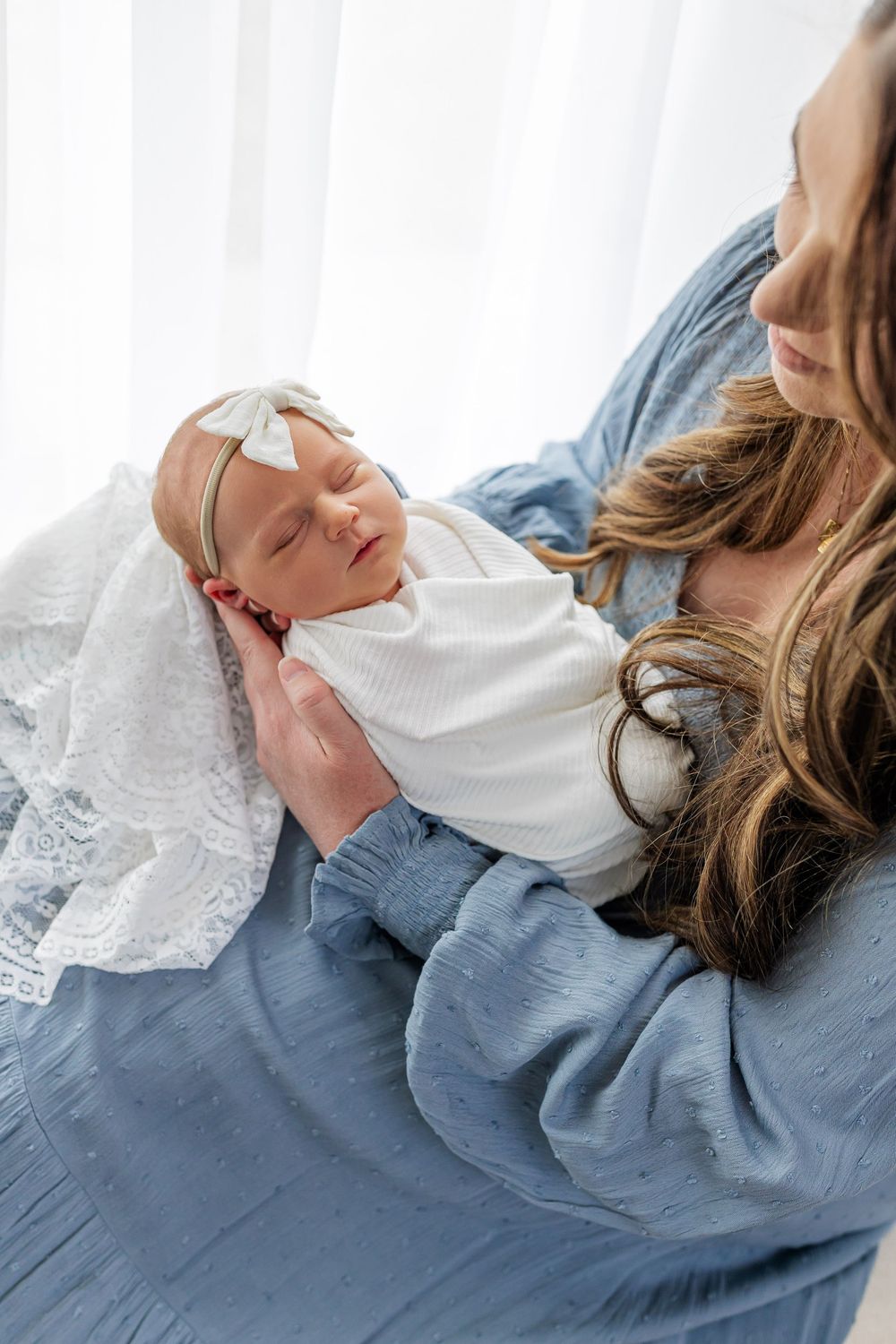 A mother in a blue dress tenderly holds her sleeping newborn wrapped in white with a bow headband near a bright window.