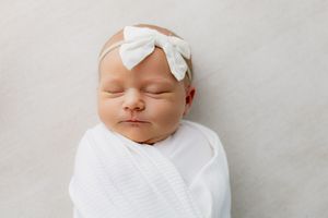 Peaceful newborn baby girl swaddled in white wrap wearing a white bow headband, sleeping on a soft white background.