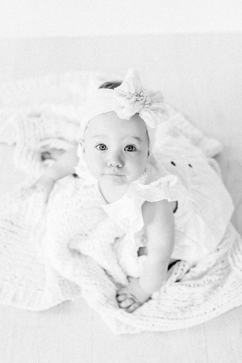A baby wearing a white dress and floral headband lies on white fabric during a lifestyle milestone session in Springfield, MO.