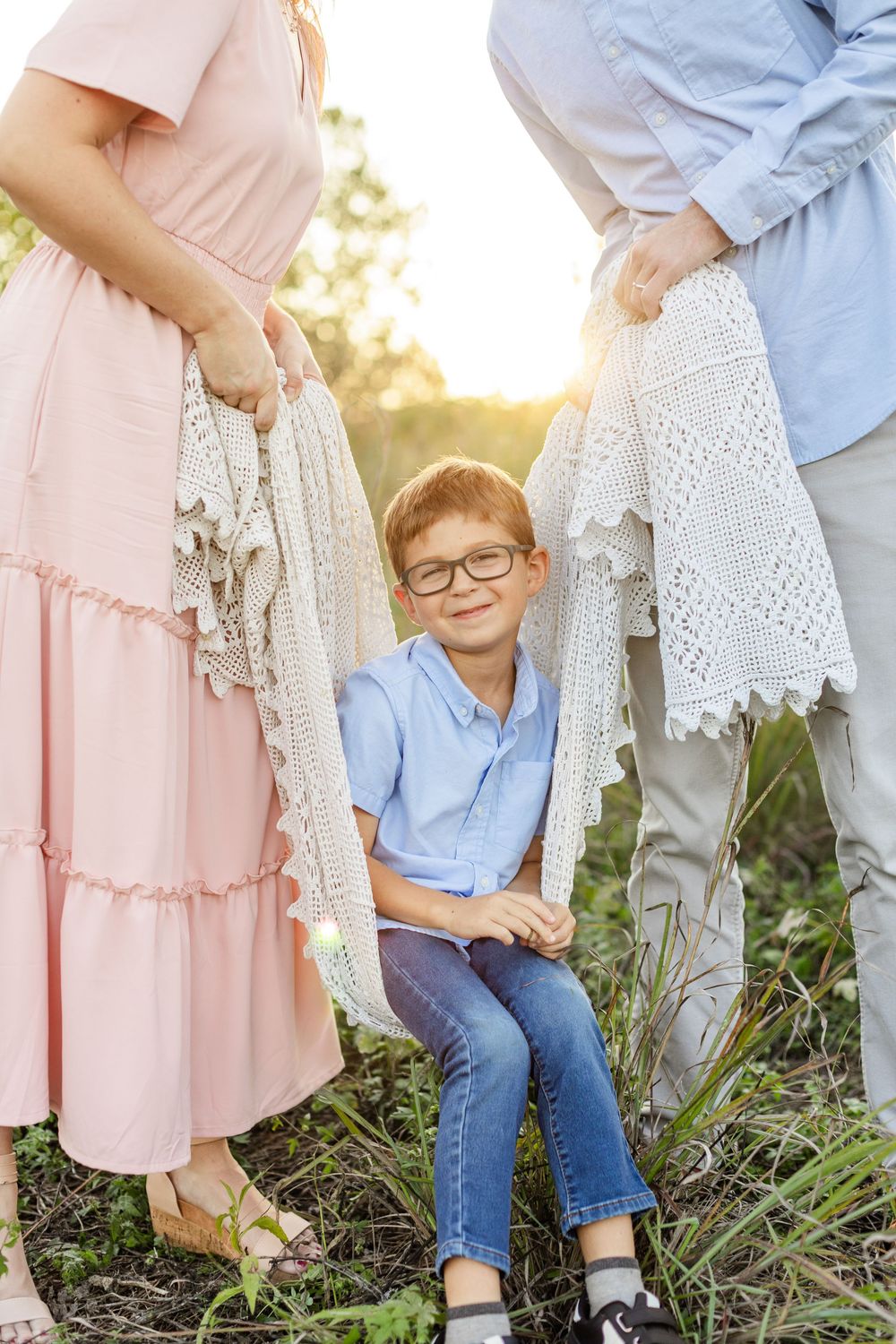 A smiling boy in glasses sits between his parents outdoors, holding a white lace blanket in golden sunset light.