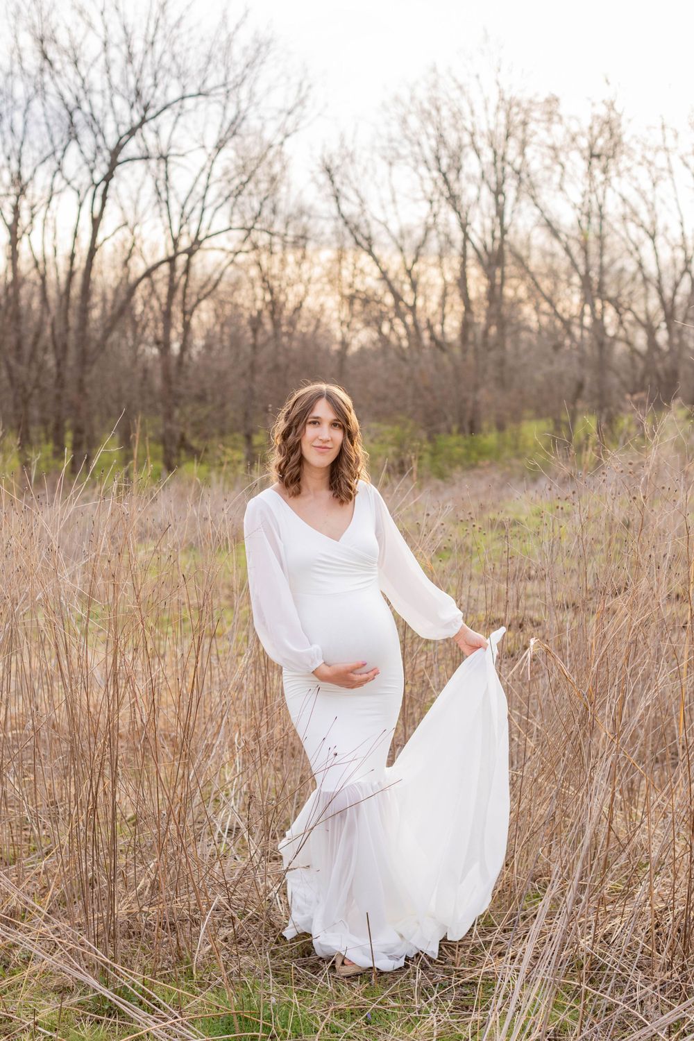 Maternity portrait of a mother in a white dress among tall grass during golden hour in Springfield, MO.