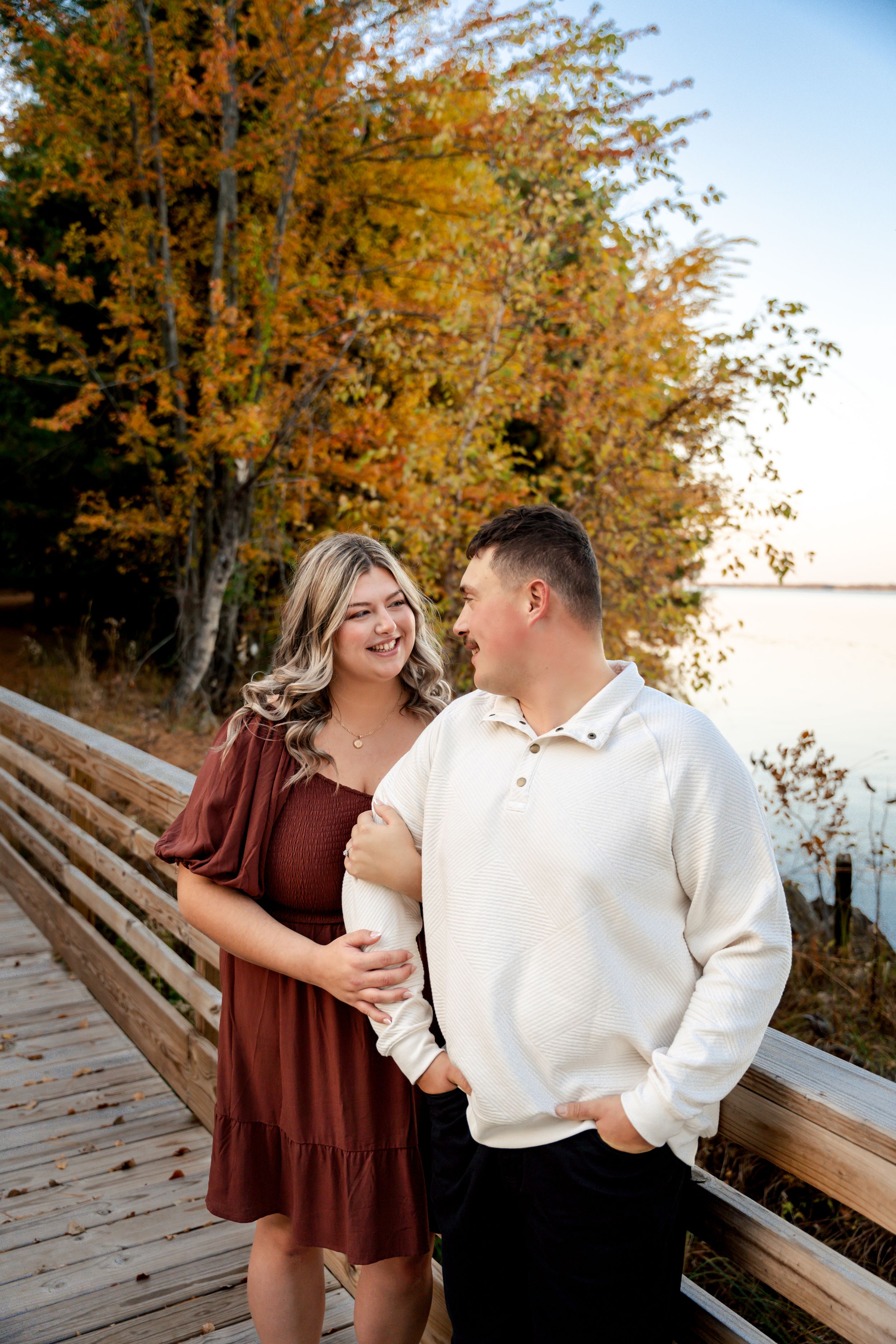 A couple shares a romantic moment on a wooden boardwalk surrounded by golden autumn foliage near a lake at sunset.