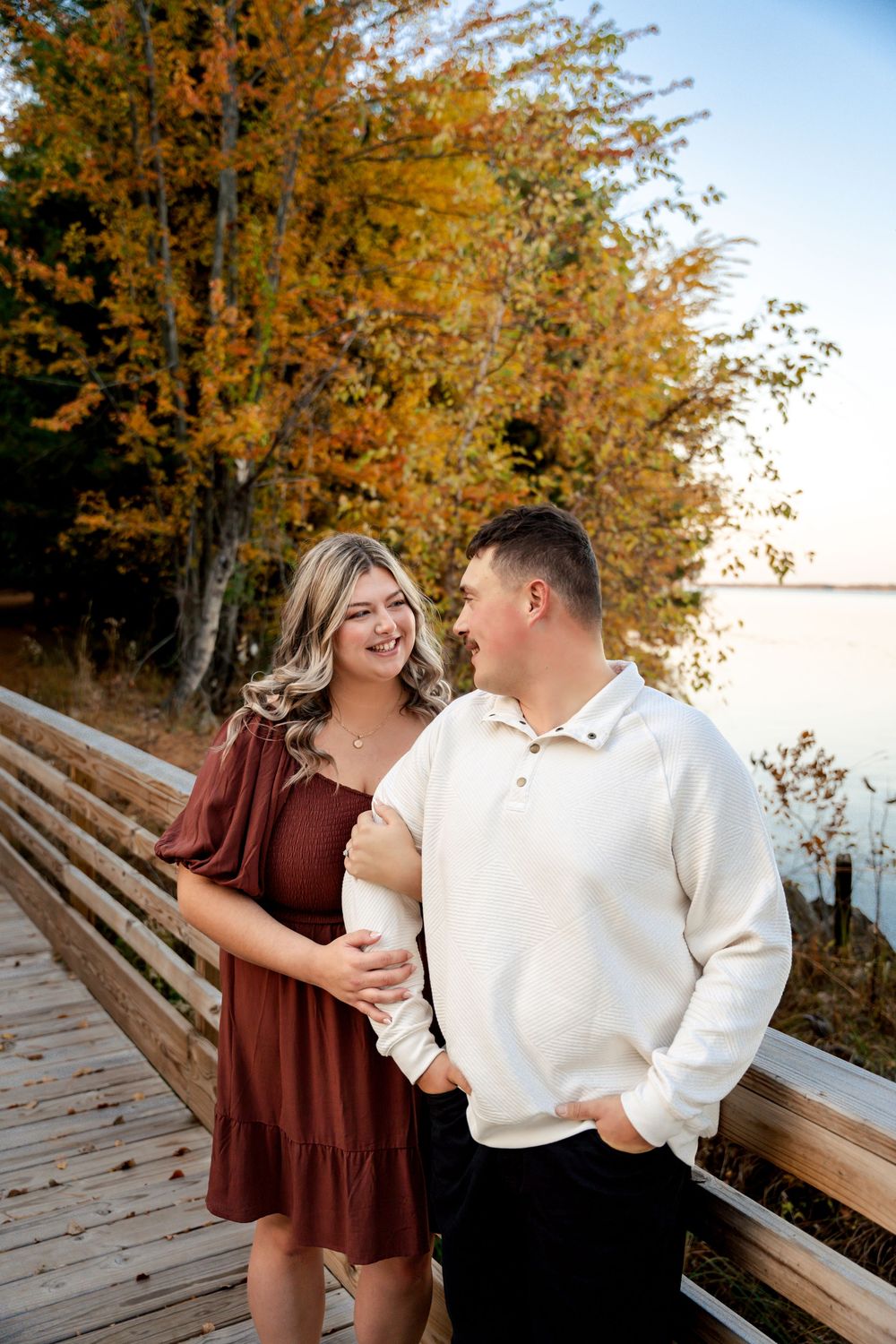 A couple shares a romantic moment on a wooden boardwalk surrounded by golden autumn foliage near a lake at sunset.