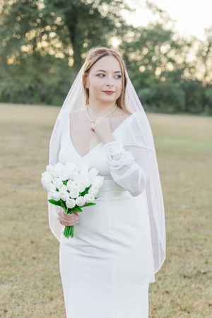 A bride in a white dress holds a bouquet of white tulips in a grassy field during golden hour.
