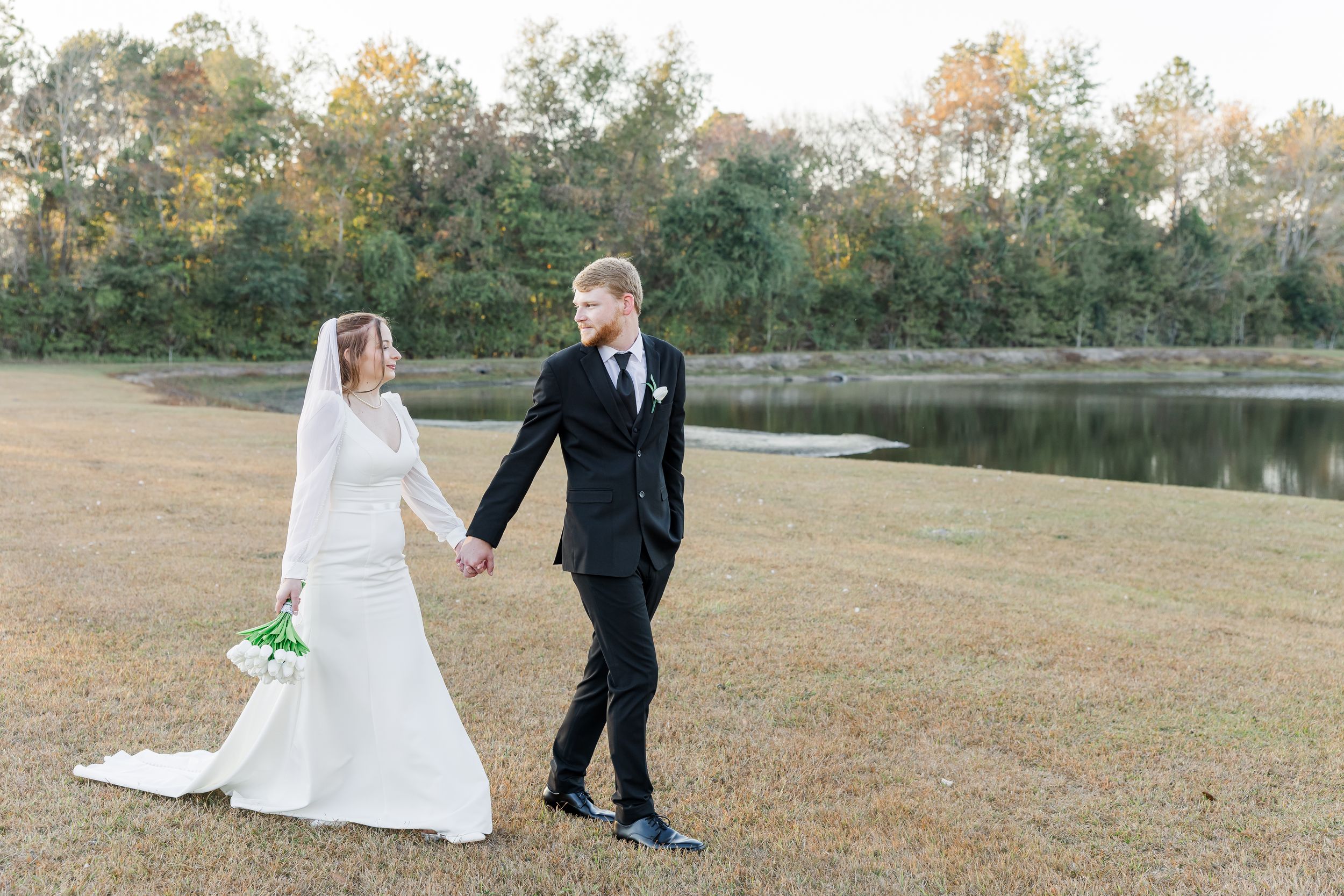 A couple in wedding attire walks hand in hand across a grassy field near a serene lake during autumn.
