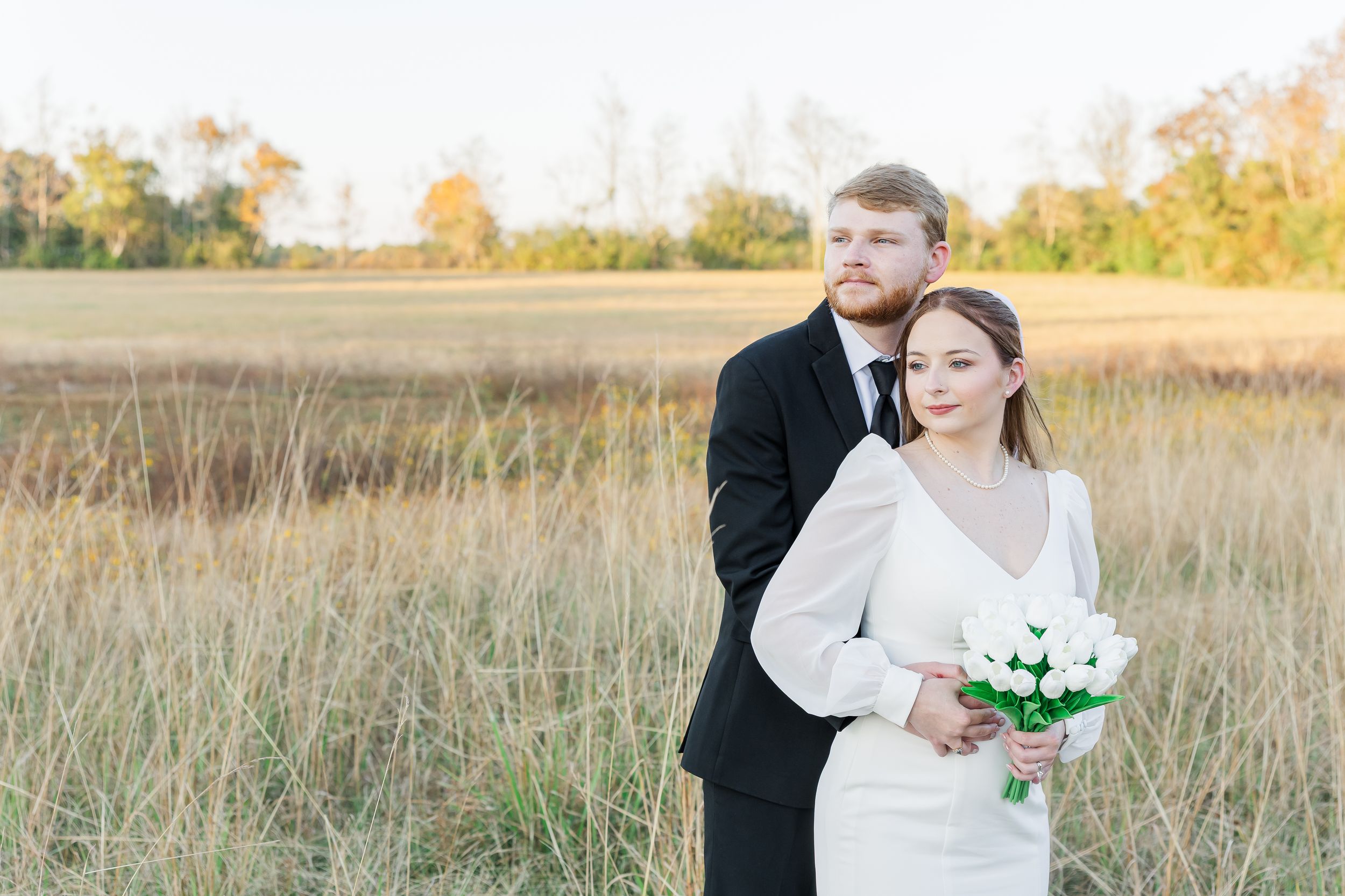 Couple in wedding attire embraces in a golden autumn field at sunset with tall grass swaying in the breeze.