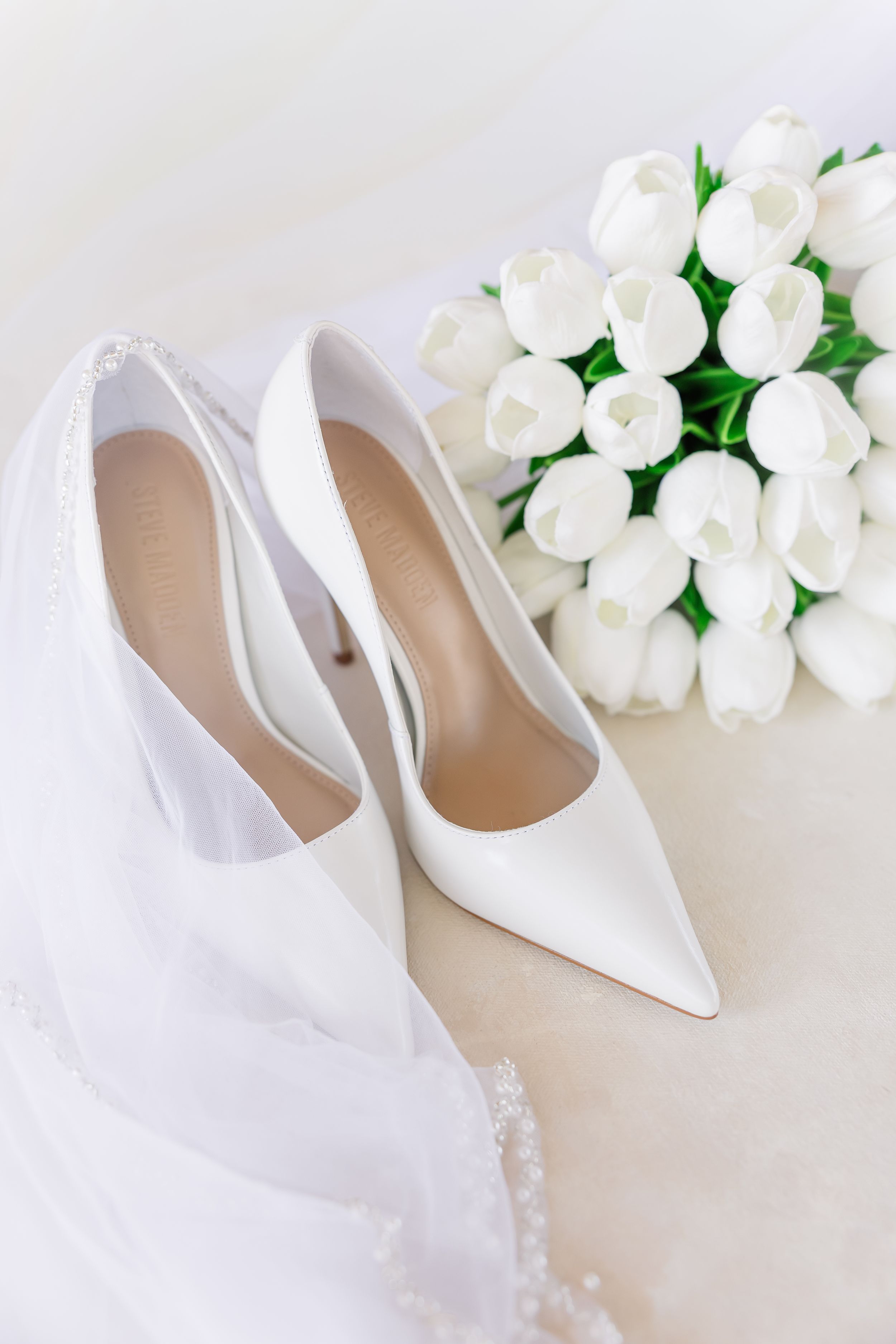 Elegant white bridal shoes with pointed toes displayed next to delicate white roses and green foliage on neutral background.