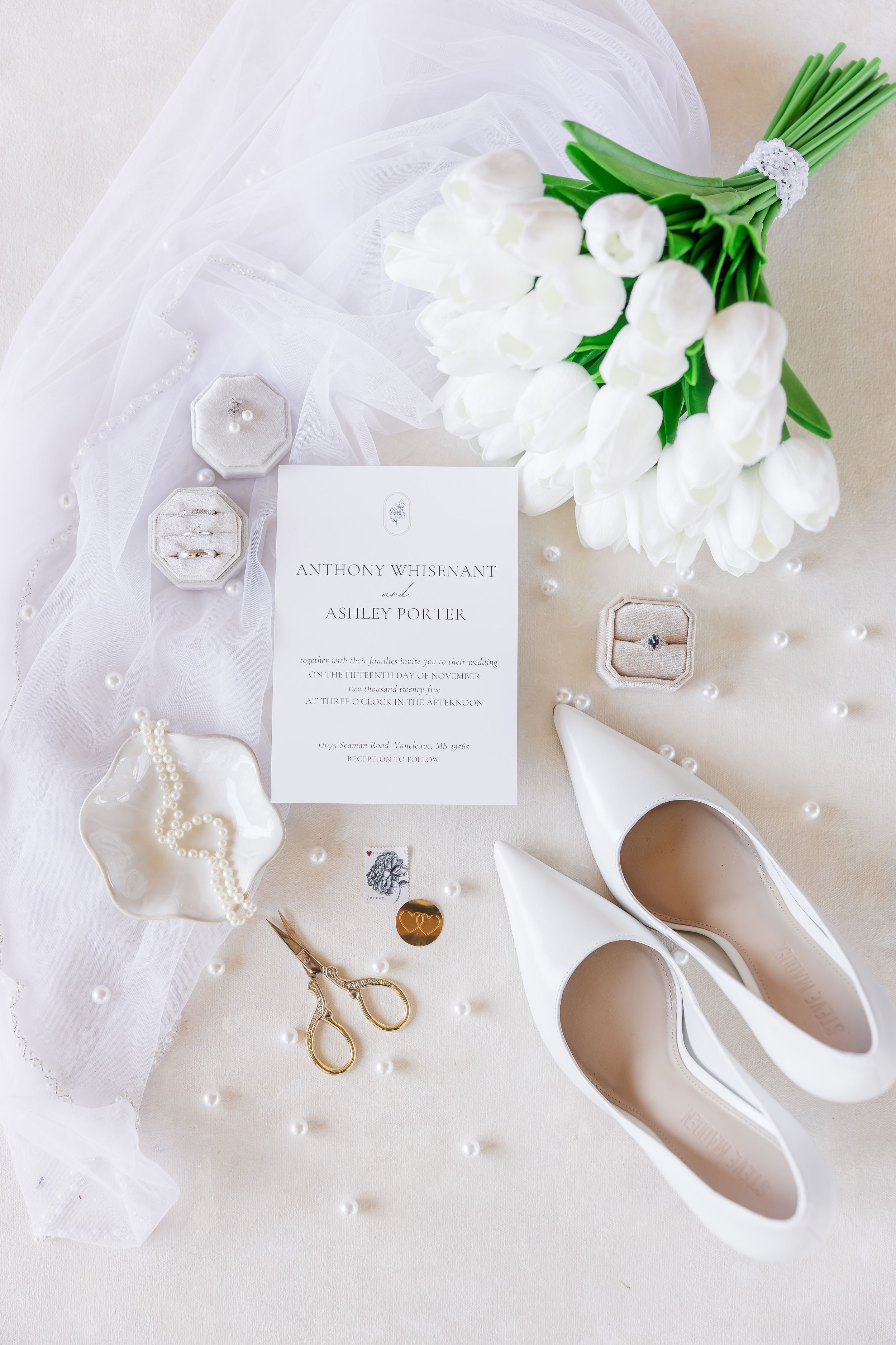 Elegant wedding details in white including shoes, flowers, invitation, and pearls arranged on a light background.