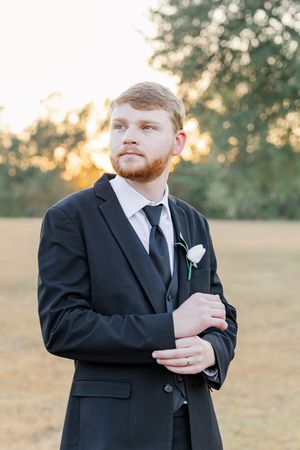 A groom in a classic black suit and tie adjusts his boutonniere in a sunlit outdoor field at golden hour.