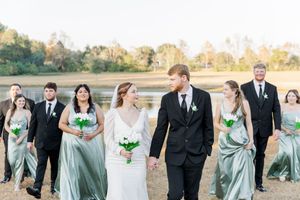 Wedding party in sage green dresses and black suits walk together on a beach at sunset.