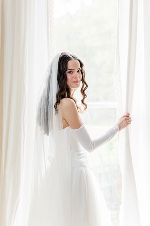 A bride in a white wedding dress and veil poses gracefully near sheer curtains in a bright sunlit room.