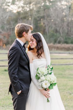 Romantic wedding couple embraces outdoors in a natural setting with white floral bouquet.
