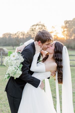 Romantic wedding couple shares a kiss at sunset in a pastoral field setting with white floral bouquet and flowing veil.