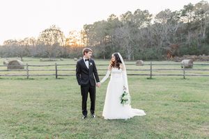 A couple in formal wedding attire walks together across a scenic grassy field during sunset.