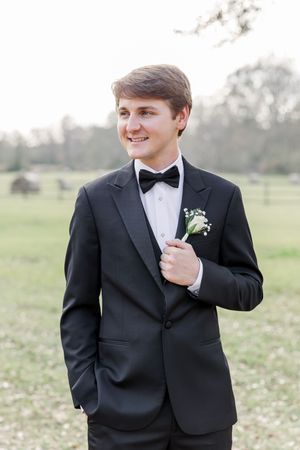 A groom in a classic black tuxedo and bowtie poses in an outdoor field during a wedding photoshoot.