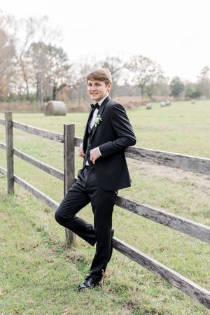 A person in a black tuxedo poses casually on a rustic wooden fence in a pastoral countryside setting.