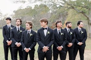 Groomsmen in black suits and bow ties stand in a row outdoors and lean forward in a synchronized pose for a fun photo.