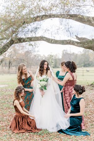 Bridesmaids in brown and teal dresses gather around a bride's flowing white dress in an autumn outdoor setting.
