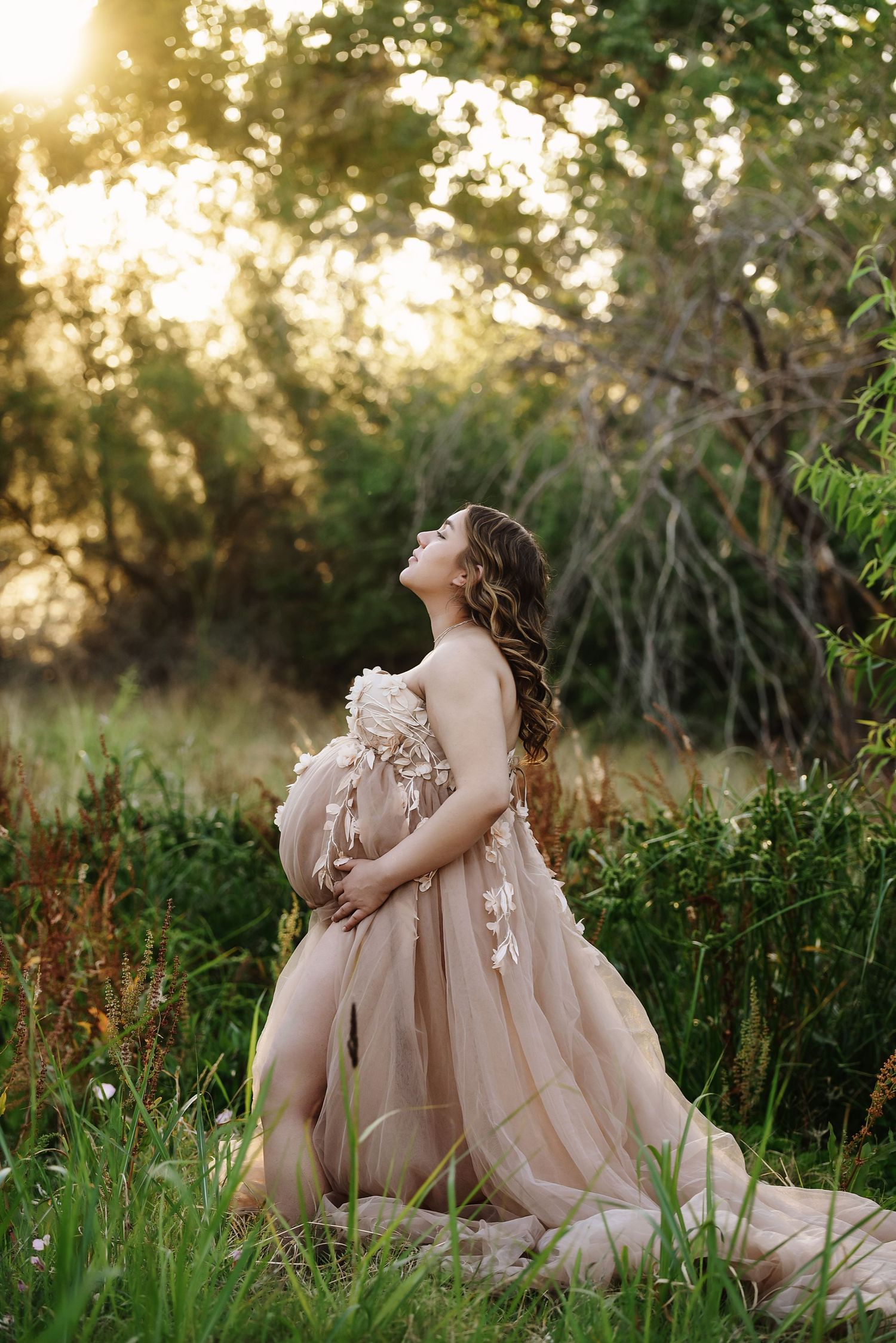 Pregnant woman in floral maternity gown standing in shallow stream surrounded by lush greenery.