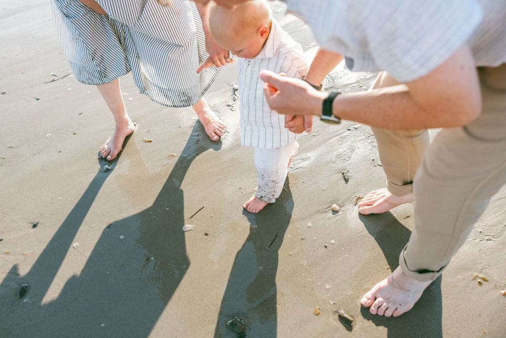 Parents helping a baby take first steps on a sandy beach, captured from above showing bare feet and shadows.