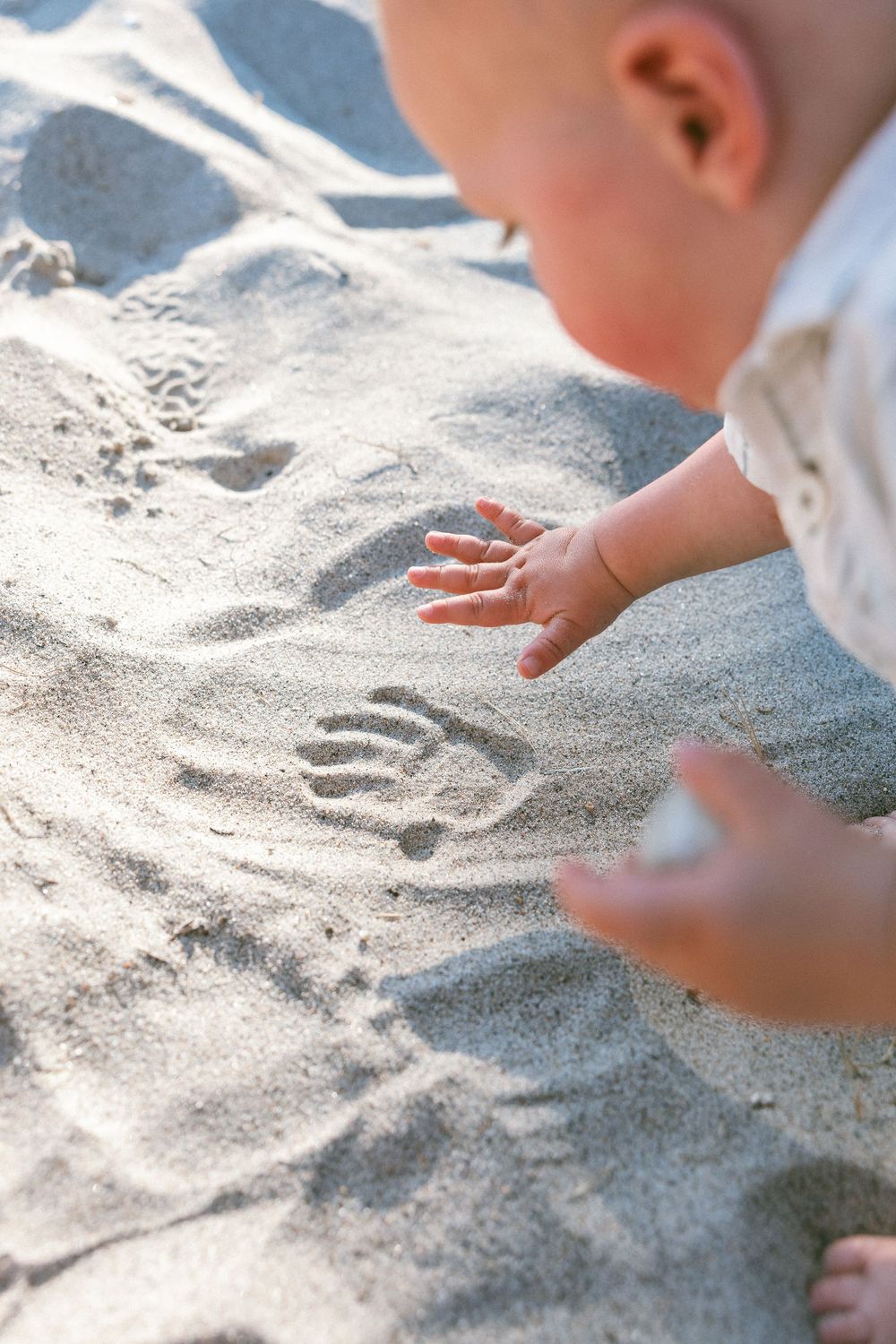 Baby's hand next to handprint pressed into sandy beach, close-up of infant exploring sand outdoors.