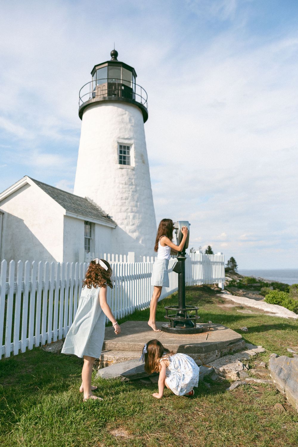 Three girls in white dresses explore a classic white lighthouse with a picket fence on a sunny day.