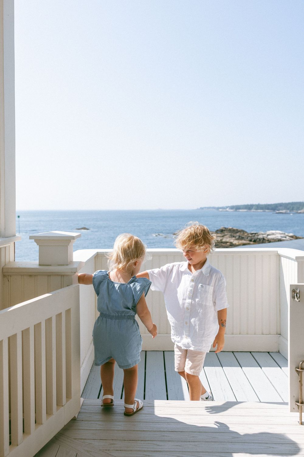 Two children in summer outfits looking out over the ocean from a white deck railing on a sunny day.