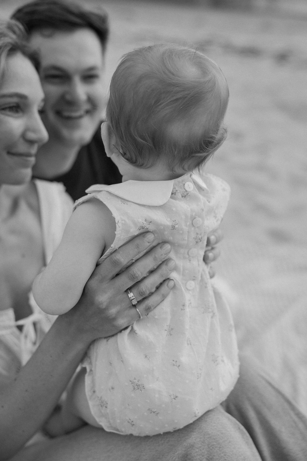 Black and white photo of smiling parents holding their baby on a beach, mother wearing a wedding ring.