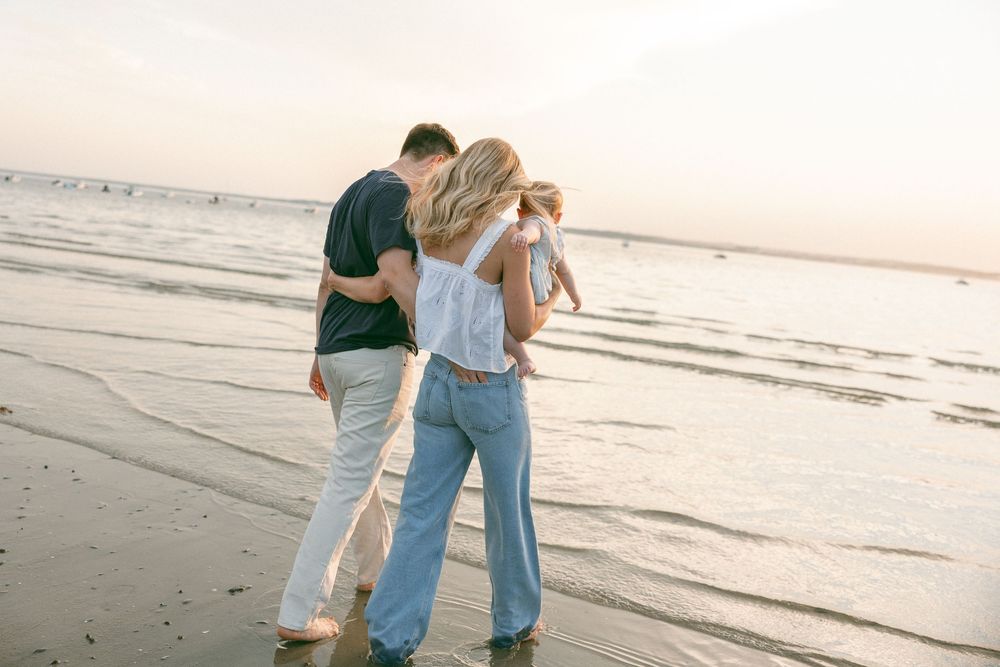 Family walking on beach at sunset, parents holding child while waves reflect golden light.