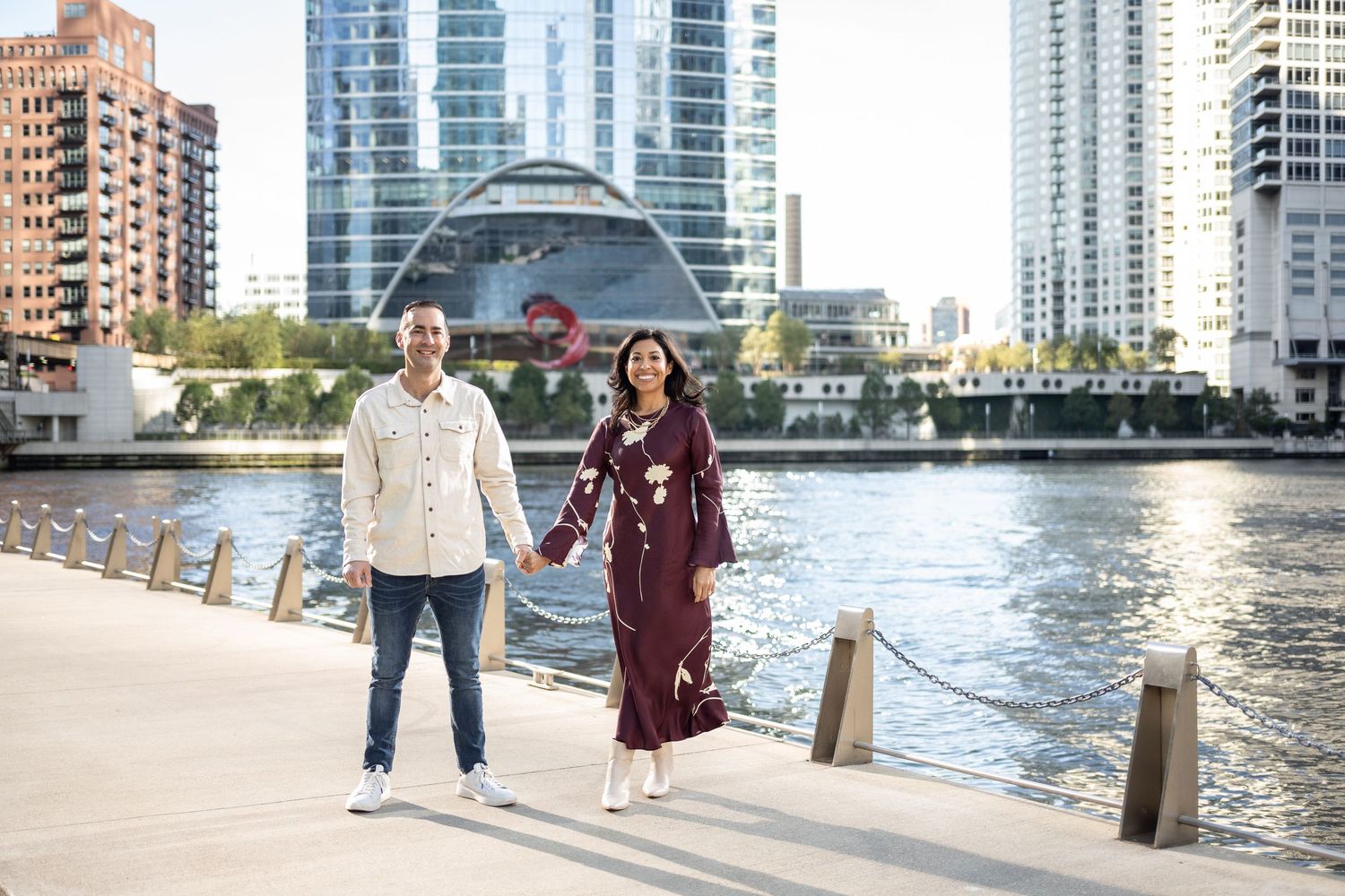 Couple holding hands on waterfront promenade with modern city skyline and glass dome building in background.