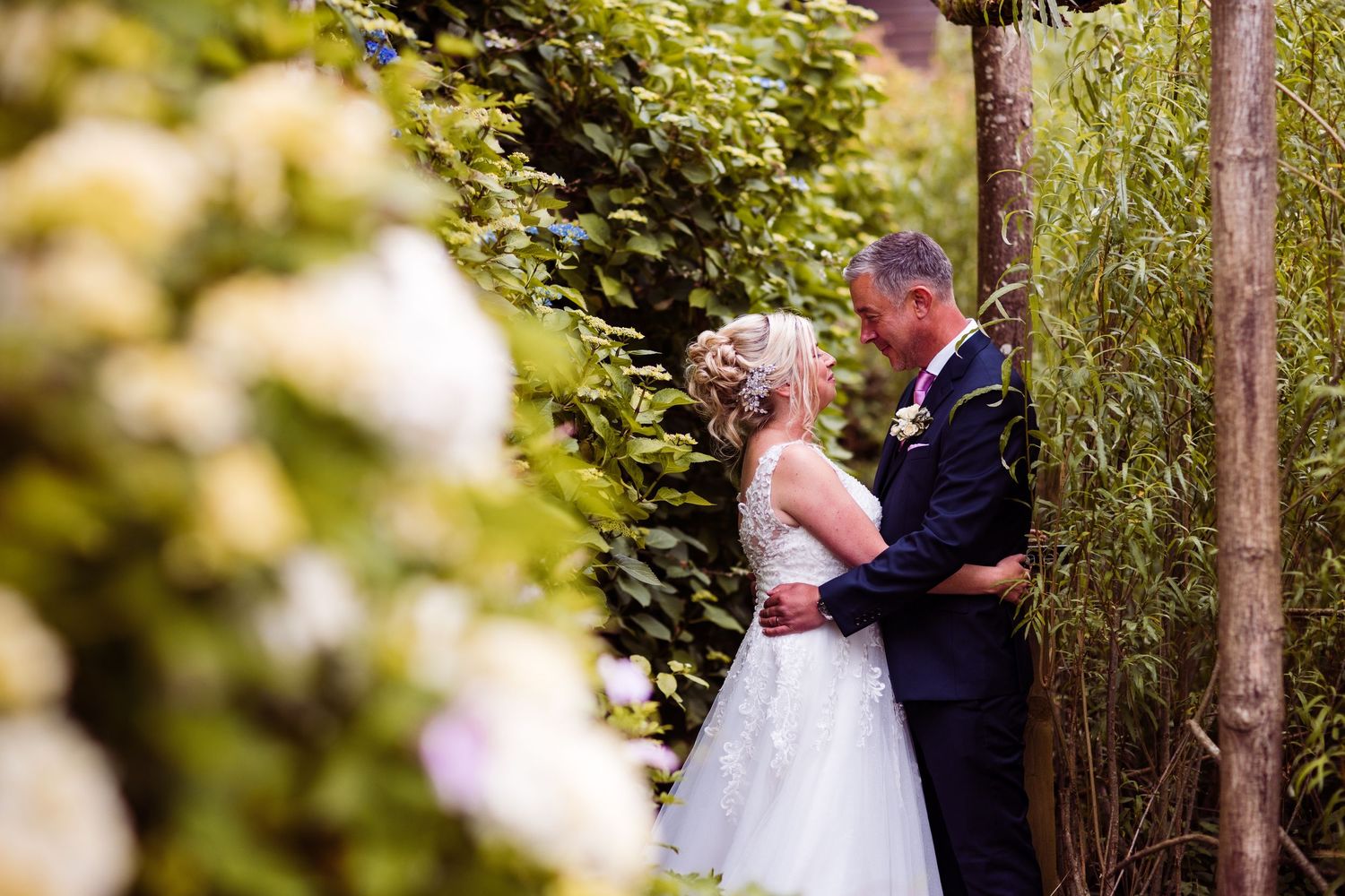 A bride and groom share a romantic moment surrounded by lush green hedges and white hydrangeas in a garden setting.