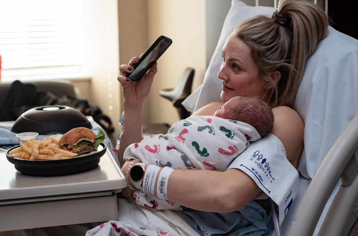 Patient relaxing in hospital bed while holding phone and having meal on tray table.
